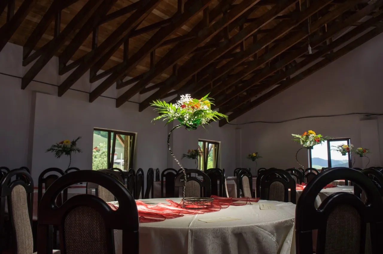 Sala de evenimente sub tavanul de bârne, cu o masă rotundă pregătită și aranjament floral înalt.