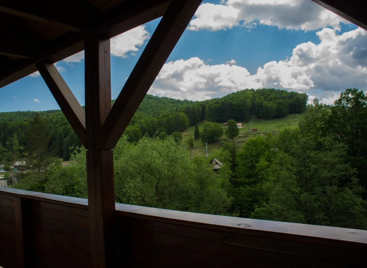 Dealurile împădurite văzute de pe balconul acoperit al cabanei, printre stâlpii de lemn.