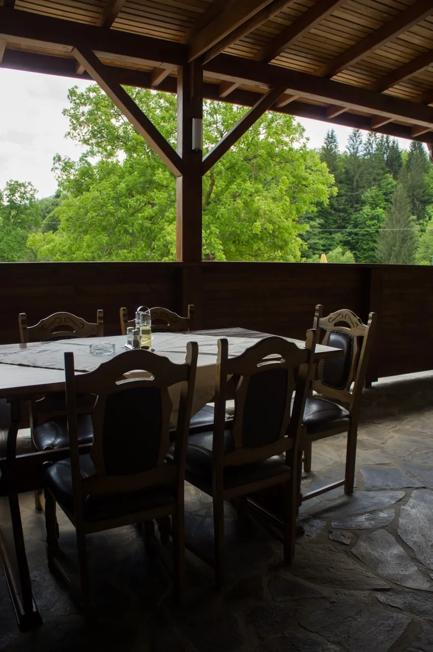 Masă de restaurant pregătită pe terasa acoperită, cu scaune de lemn și verdeață în spate.
