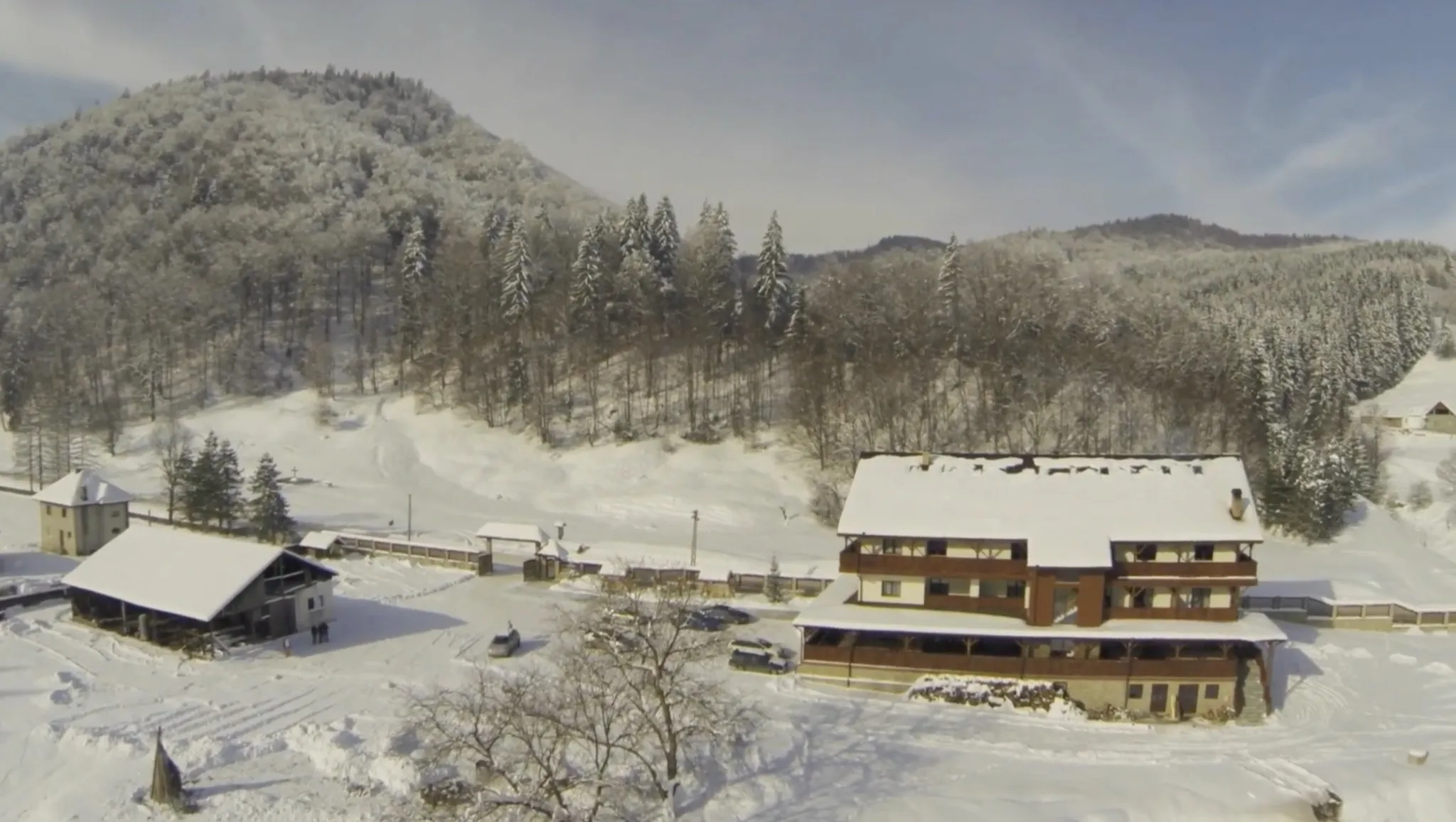 Vedere aeriană largă, din spate: cabana în dreapta, o anexă în stânga, curtea între ele, iar dealul de brad și fag umple jumătatea de sus a cadrului.