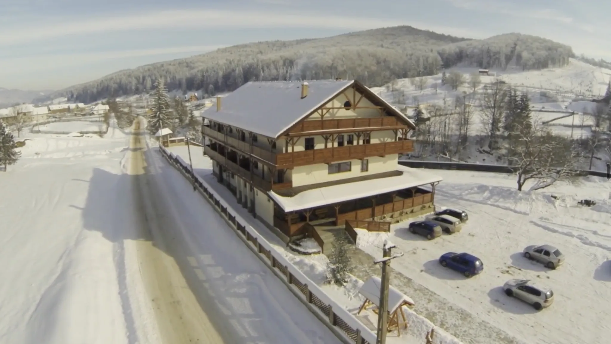Vedere aeriană de iarnă: cabana întreagă dinspre drum, cu zăpadă pe acoperiș, drumul deszăpezit și gardul lui, curtea cu mașini parcate și dealurile împădurite în spate.