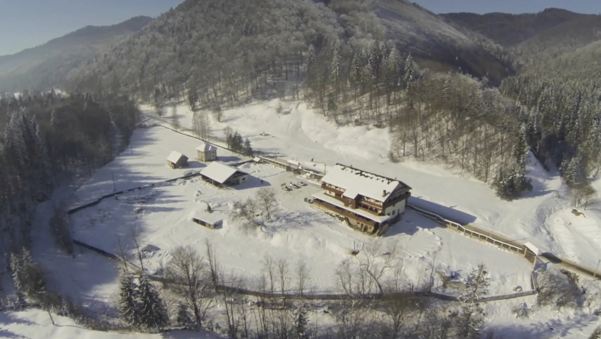 Vedere aeriană de sus, iarna: cabana, anexele, curtea și gardurile ei în vale, înconjurate de dealuri împădurite și înzăpezite.