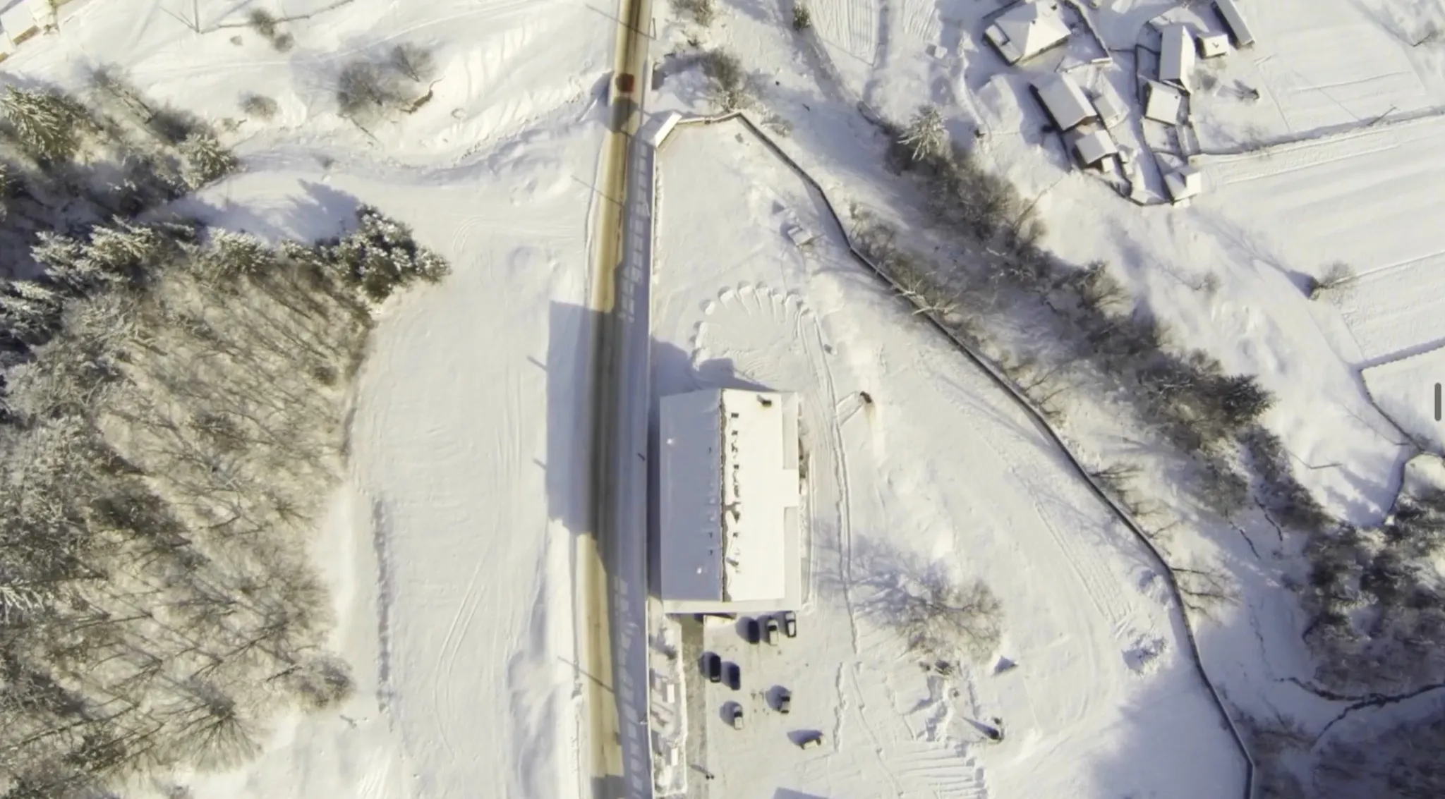 Vedere aeriană verticală, de deasupra: acoperișul cabanei acoperit de zăpadă, drumul deszăpezit care trece pe lângă el, mașinile parcate și acoperișurile vecinilor în dreapta sus.