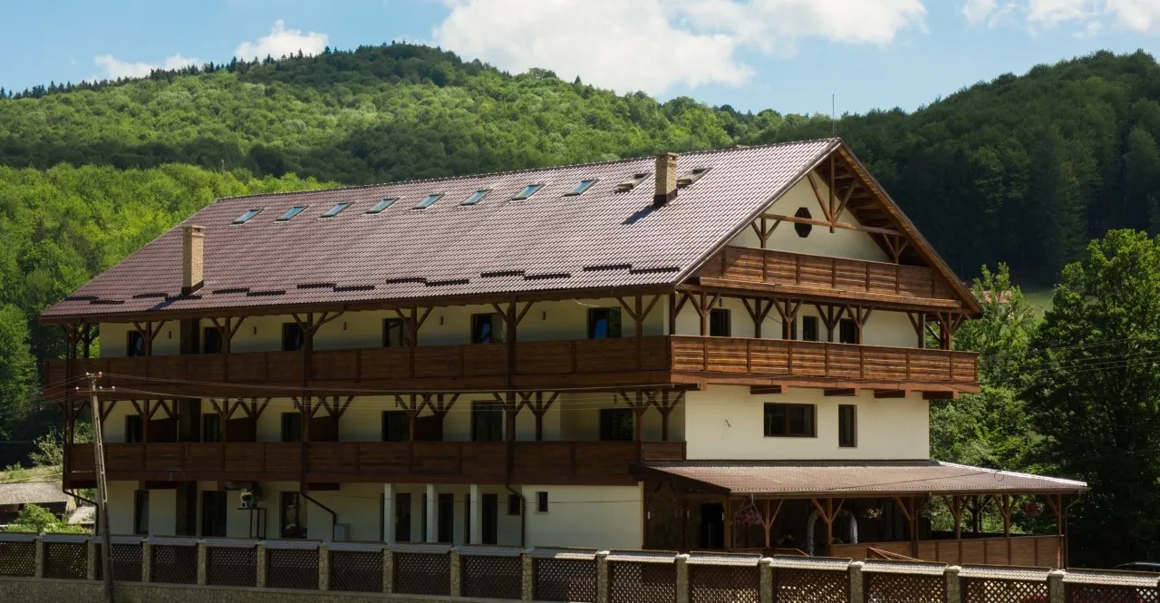 Cabana văzută pe diagonală, cu balcoane de lemn pe trei niveluri de-a lungul fațadei și frontonul acoperișului la capăt, dealul împădurit în spate.