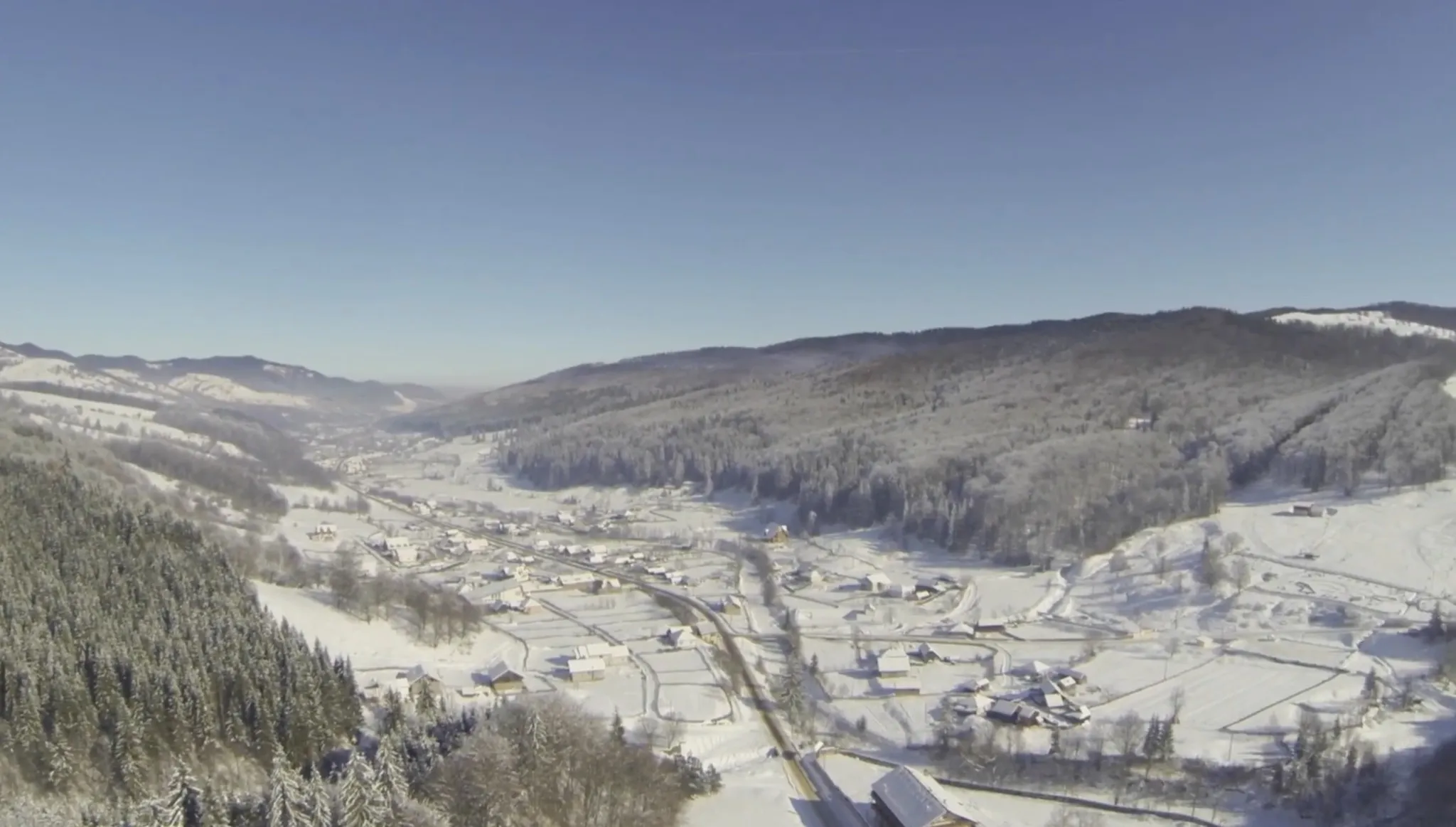 Vedere aeriană de sus, în lungul văii: satul înșirat pe lângă drum, parcele de câmp înzăpezite, creste împădurite pe amândouă laturile și cer albastru. Cabana nu se distinge în acest cadru.