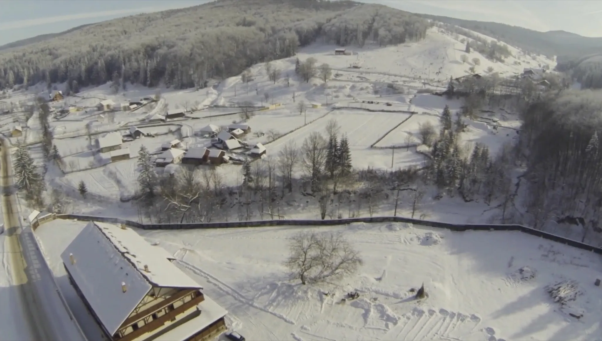 Vedere aeriană de deasupra acoperișului cabanei — care se vede în stânga jos — spre satul Văleni-Stânișoara: parcele îngrădite, case și dealul împădurit în spate.